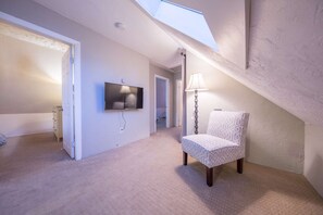 A spacious room with a sloped ceiling, a flat-screen TV mounted on the wall, a patterned armchair, and a floor lamp. - Riverview (Beverly)