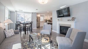 A modern living room with a sofa, armchairs, a coffee table, and a fireplace. There is a dining area with a table and chairs, and a kitchen in the background.