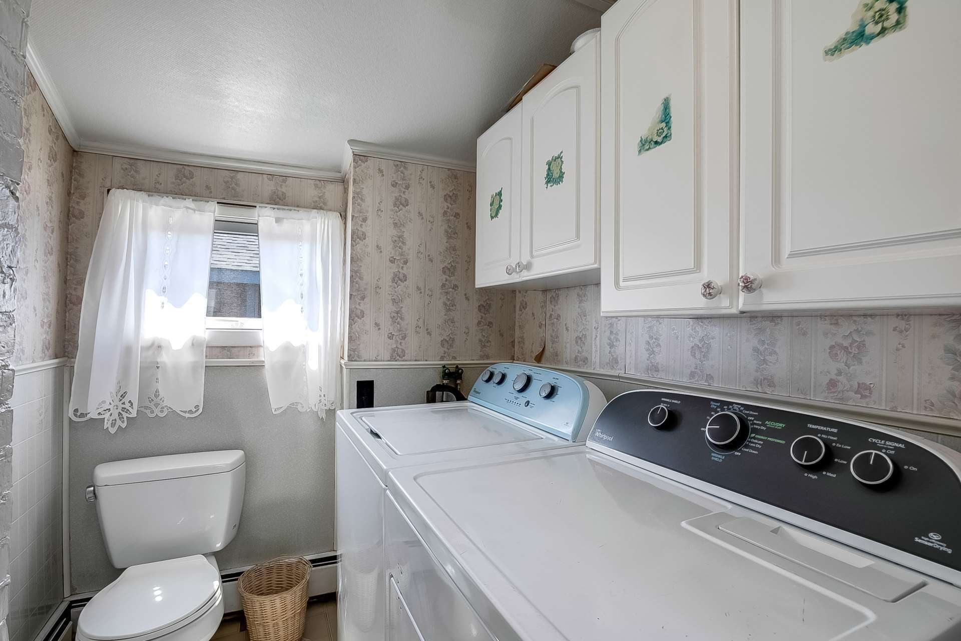 A compact laundry room with a front-loading washer and dryer, a toilet, and a window with sheer curtains.