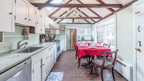 A kitchen with a dining area, featuring a brick floor, white cabinets, a red tablecloth-covered table, and a chandelier. - Rockmere Inn (Marblehead)