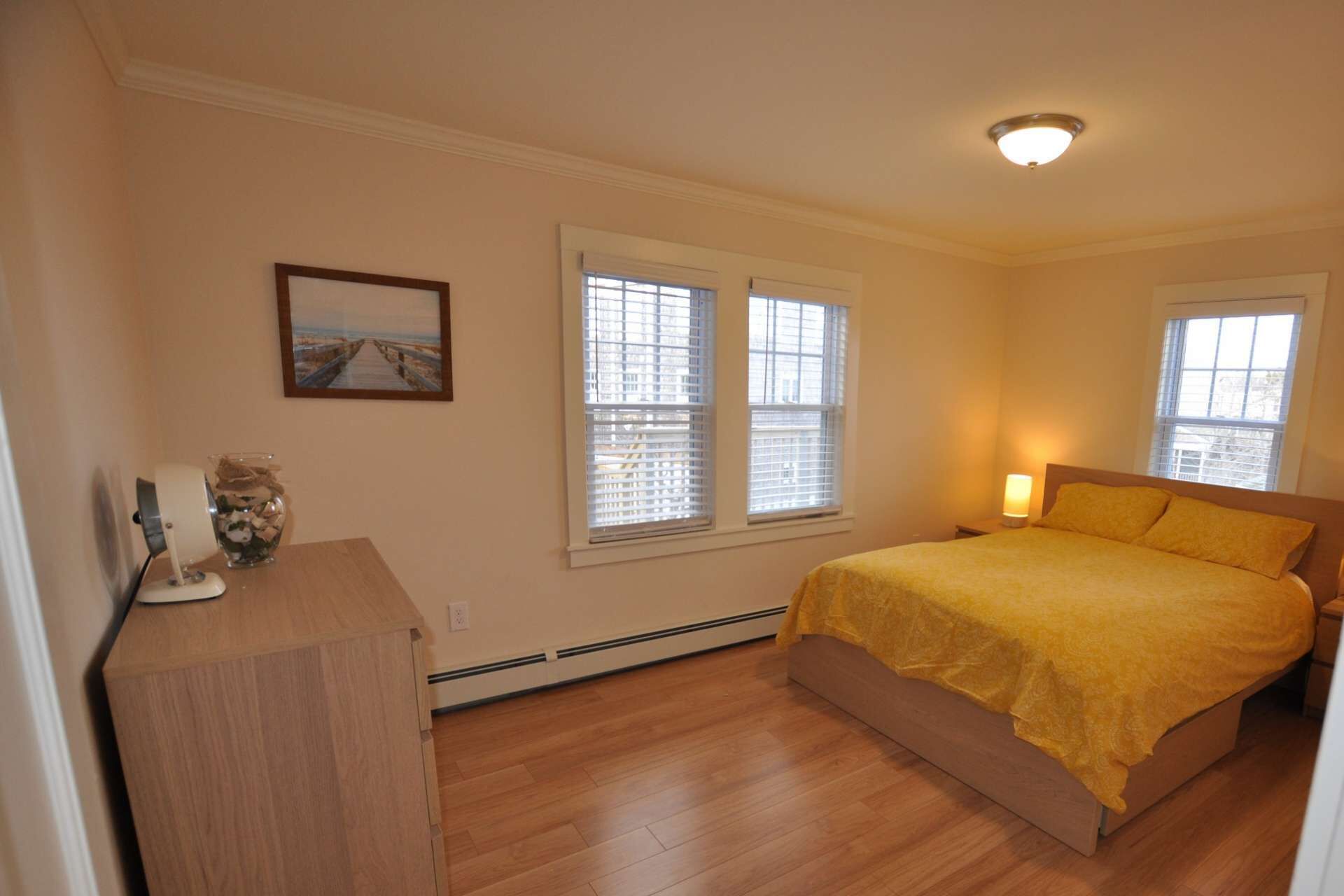 A bedroom with a bed, two windows, a picture on the wall, and a nightstand with a lamp.