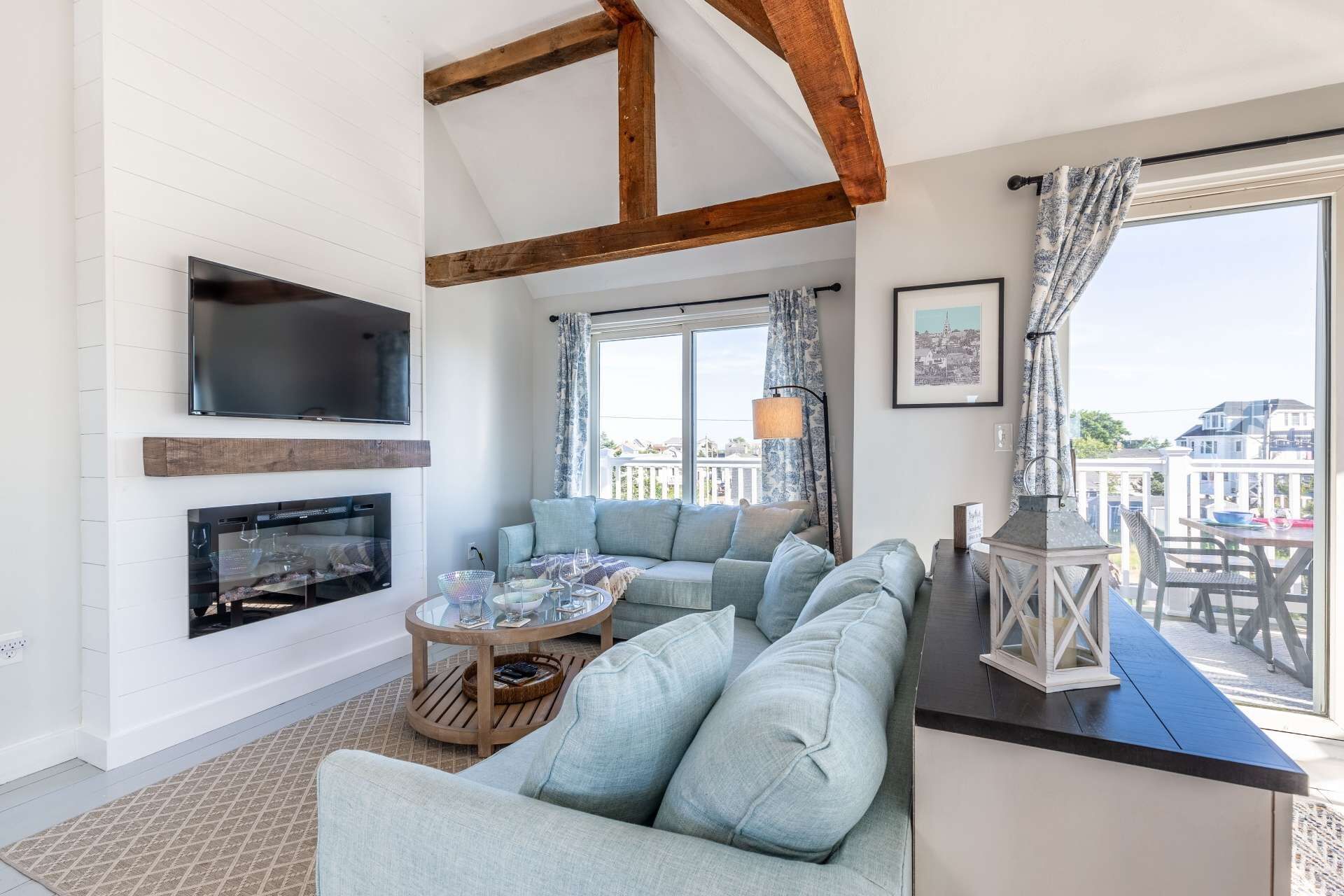 A living room with a large sofa, a television, and a fireplace. There is a balcony with a table and chairs.