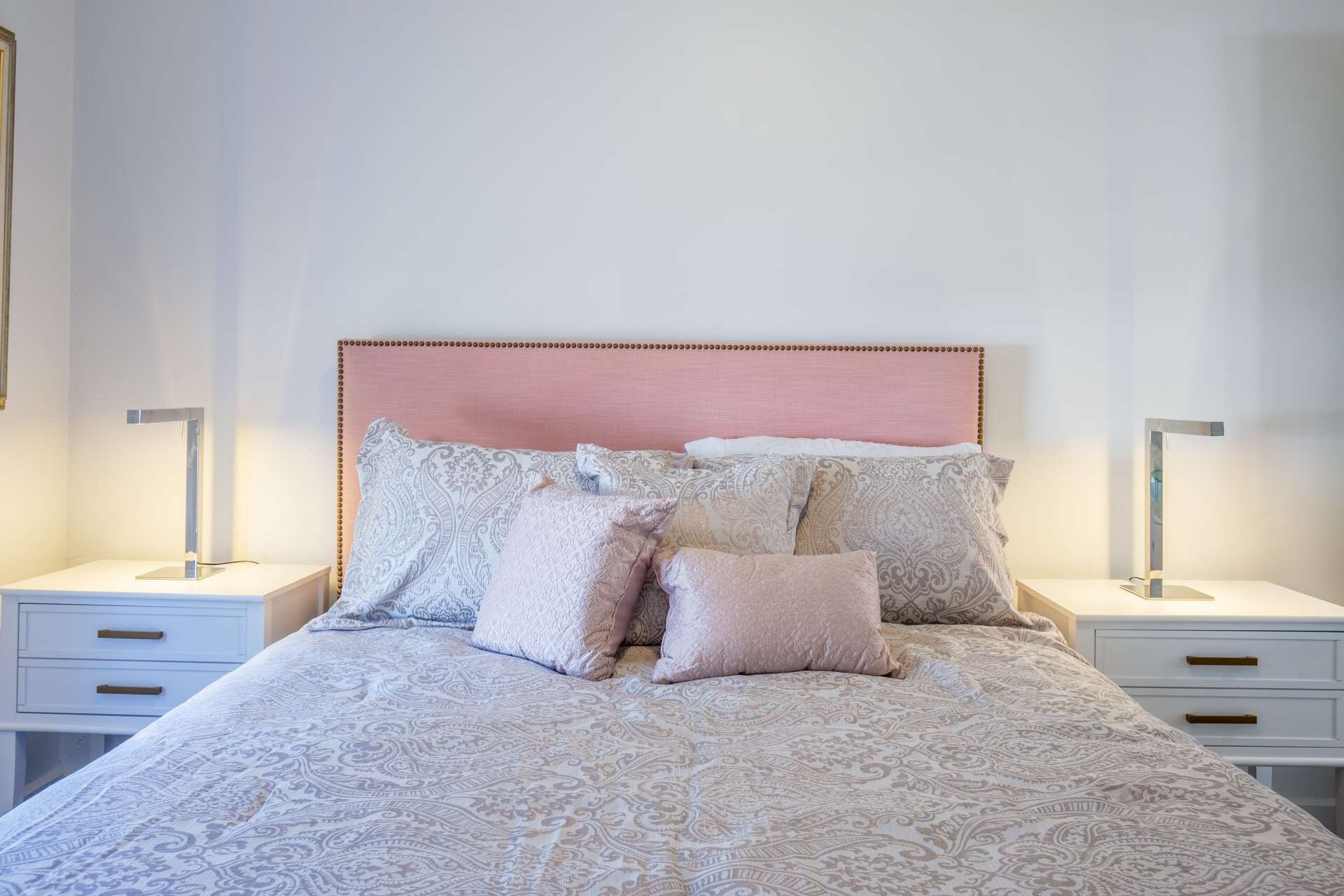 A neatly made bed with patterned bedding, a pink headboard, and bedside tables with lamps.