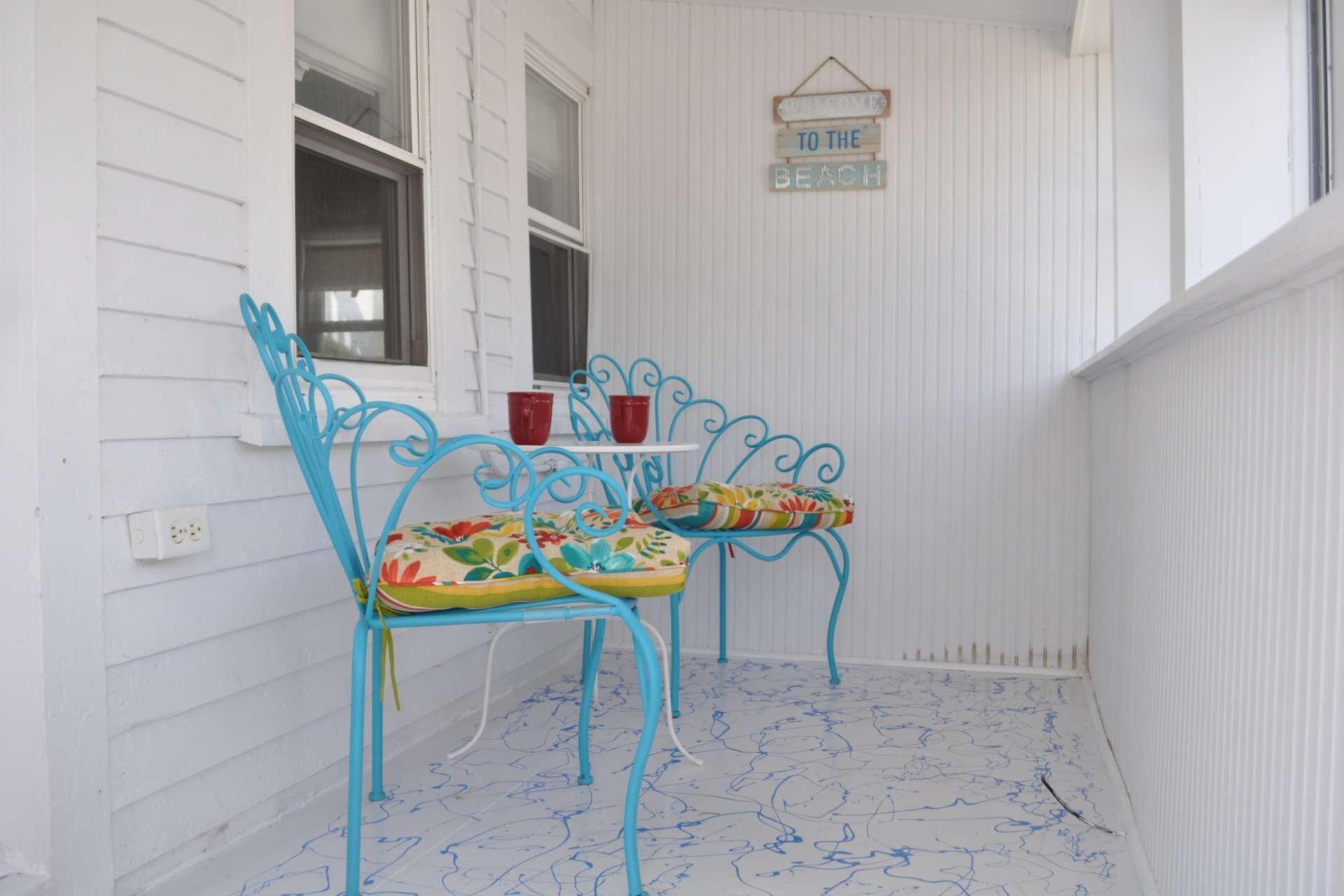 En veranda med två blå metallstolar, var och en med en färgglad kudde. Ett litet bord med en röd kopp och en vit mugg står mellan stolarna. En skylt ovanför lyder: "TILL STRANDEN".