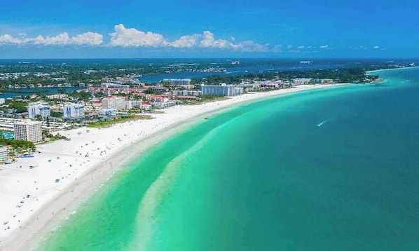Un paysage urbain côtier avec une vaste plage, des eaux turquoise et limpides, et une silhouette de bâtiments qui se dessine à l’horizon.