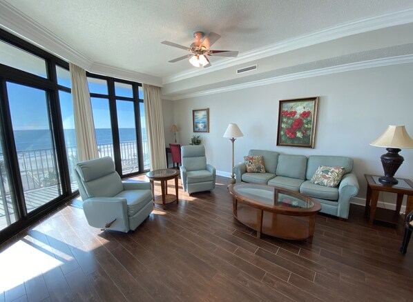 A spacious living room with a view of the ocean, featuring a ceiling fan, a light-colored sofa, two armchairs, a coffee table, and a floor lamp.