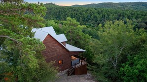 A wooden cabin with a metal roof, surrounded by dense forest, and a staircase leading to a deck. - Whitetail Ridge Has Unbelievable Views Overlooking Beavers Bend State Park (Broken Bow)
