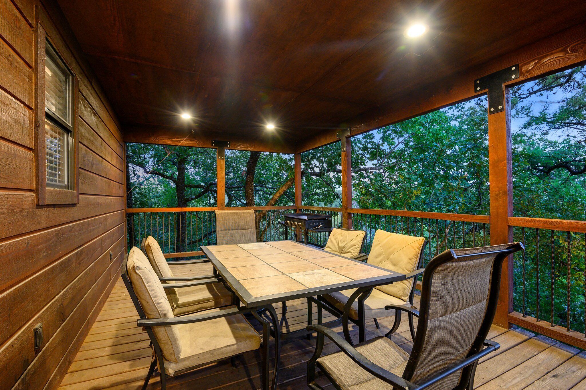A covered outdoor patio with a wooden table and chairs, surrounded by a wooden railing, overlooking a forested area.