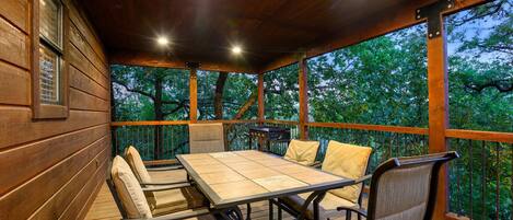 A covered outdoor patio with a wooden table and chairs, surrounded by a wooden railing, overlooking a forested area.