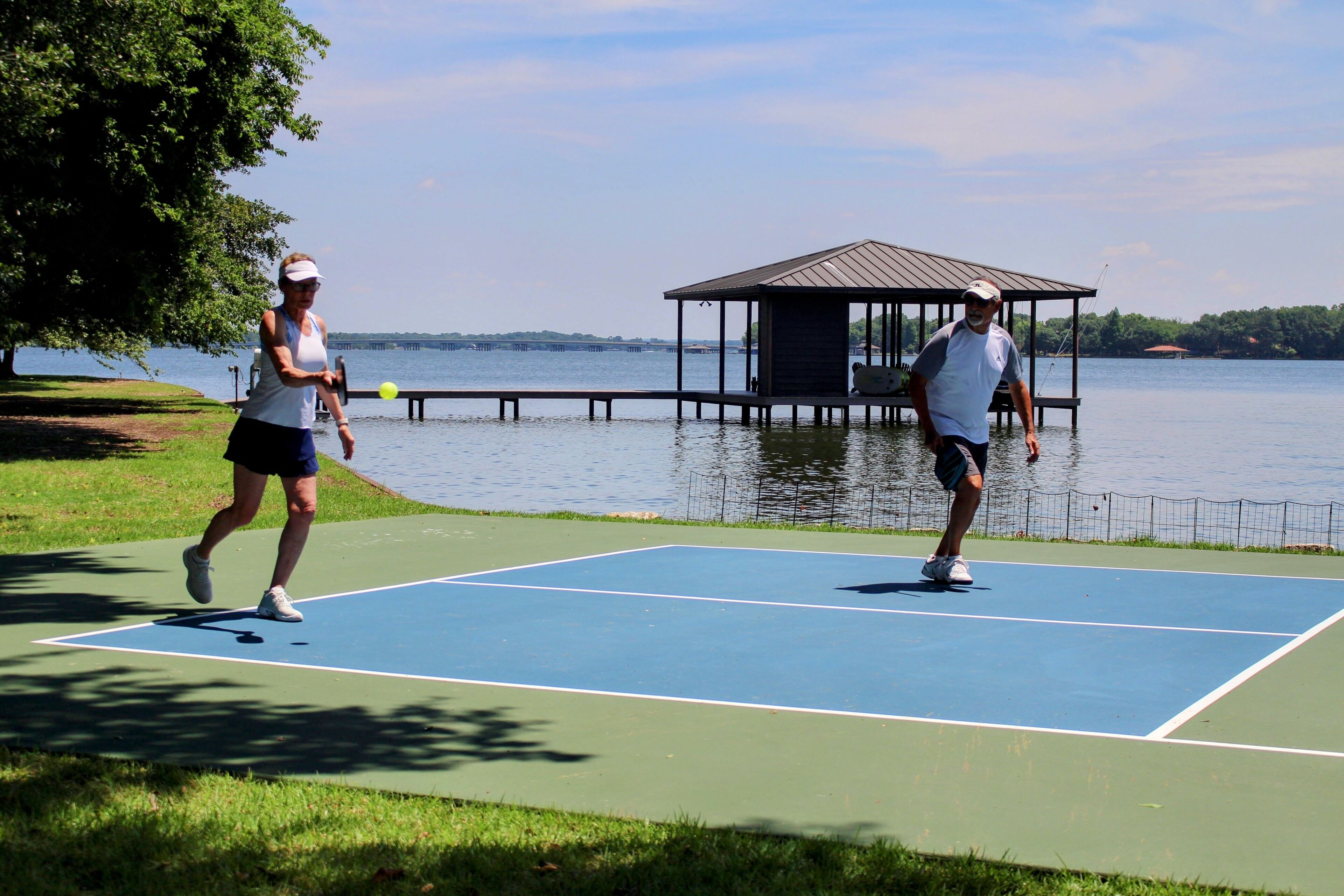 Zwei Personen spielen Tennis auf einem Platz, im Hintergrund ein See und eine Laube.