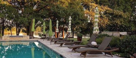Un área junto a la piscina con sillones de descanso y sombrillas, rodeada de árboles.