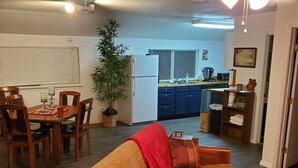 A cozy living room with a sofa, a dining table, and chairs. There is a kitchen area in the background with cabinets and a refrigerator.