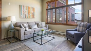 Un salon comprenant un canapé, un fauteuil, une table basse en verre et une grande fenêtre donnant sur des immeubles.