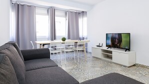 A modern living room with a grey sofa, a dining table with chairs, a TV on a white stand, and large windows with curtains. - Spacious, Central Apartment – Ideal for families (Salou)