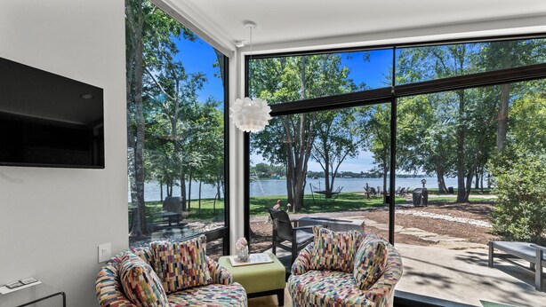 A modern living room with large windows overlooking a lake and park. There are two patterned armchairs and a small table with a vase. - Adults-Only Lake Peninsula Cottage w/ Pool, Pickleball & Sunset Views (Malakoff)