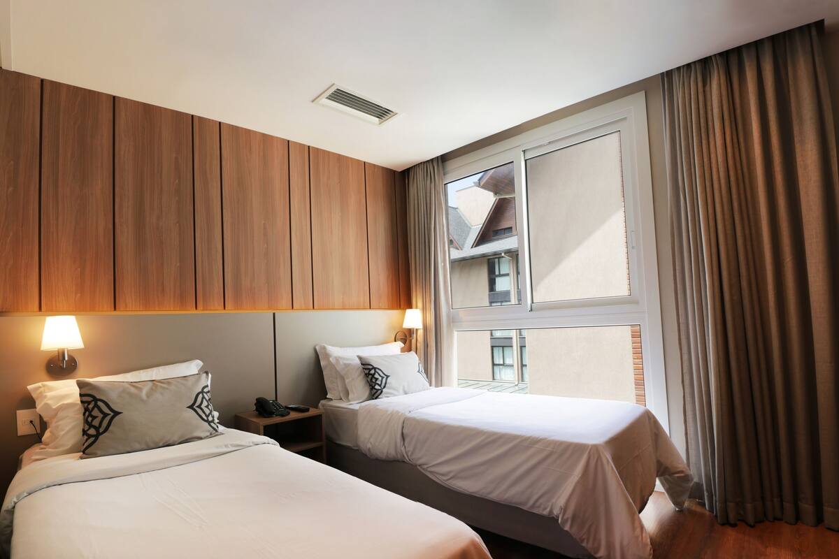 A hotel room with two beds, wooden paneling on the walls, a large window with curtains, and a visible balcony.