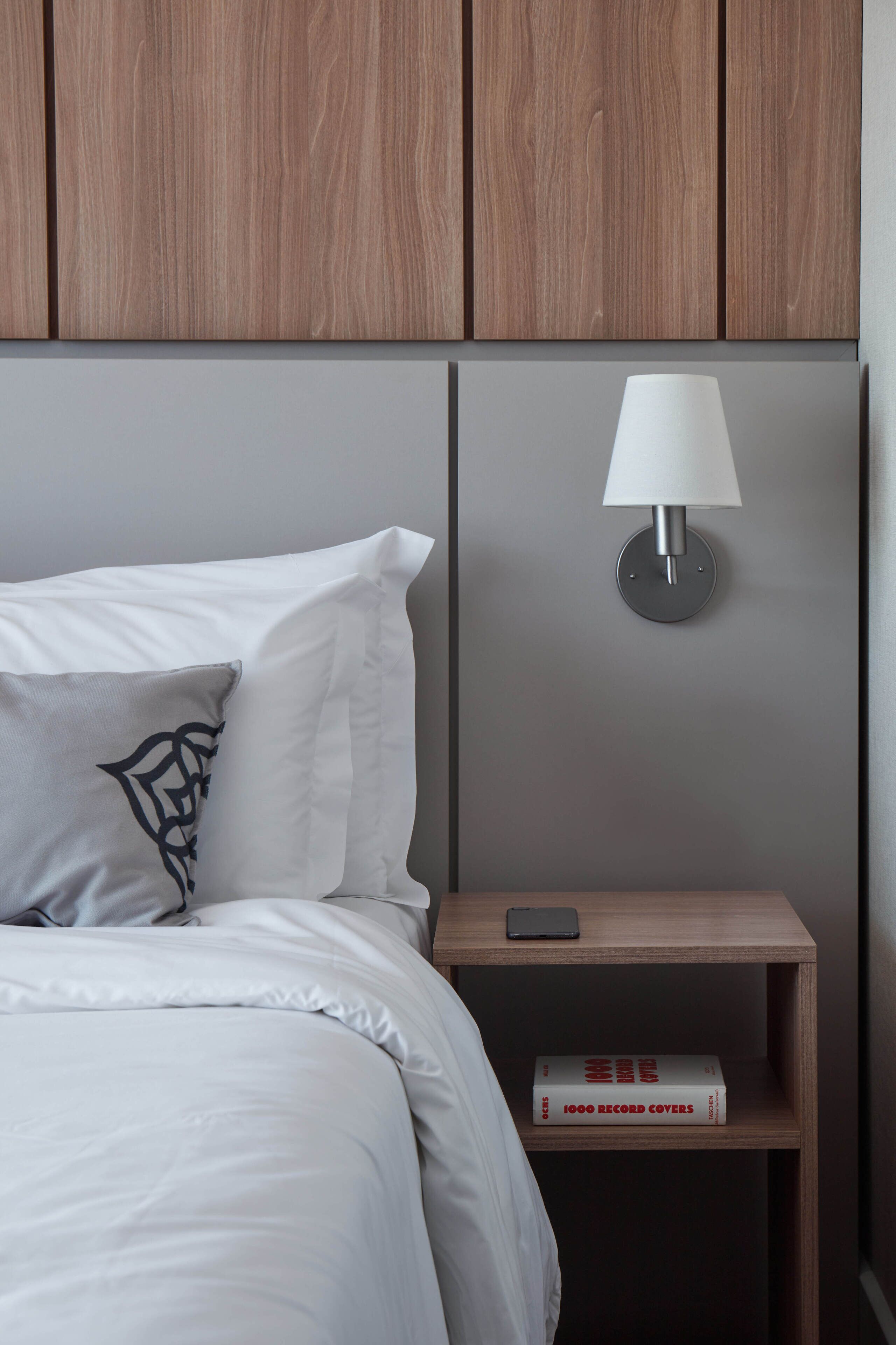 A neatly made bed with white bedding and a decorative pillow, a bedside table with a lamp and a book titled "1000 Record Covers," and wooden paneling above the bed.