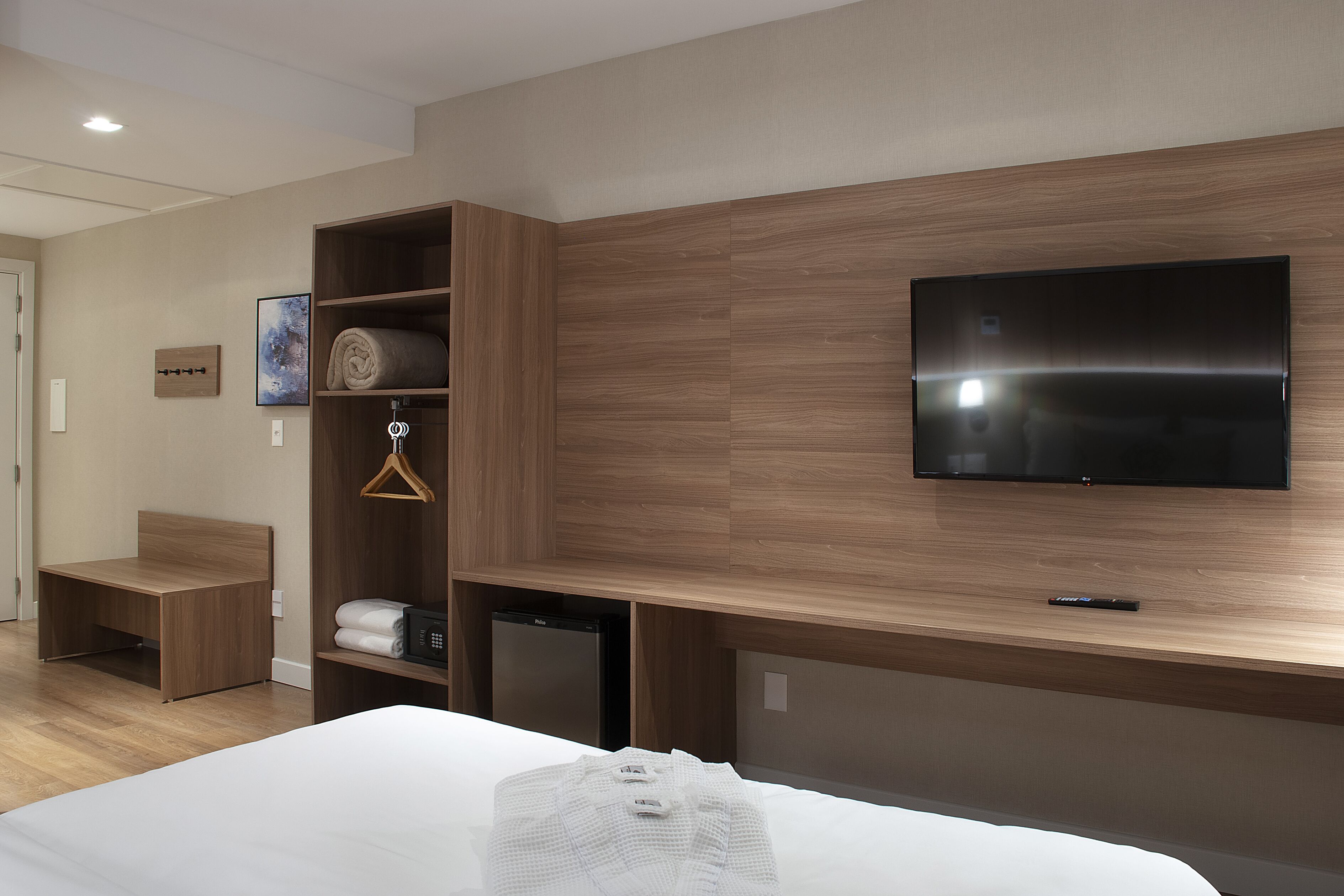 Een hotelkamer met een bed, een aan de muur bevestigde televisie, een houten garderobe, een klein bureau en een minibar.