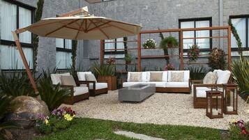 Uma área externa moderna com assento, incluindo um sofá, cadeiras e uma mesa de centro. Há uma pérgola de madeira com vasos suspensos e um guarda-sol grande.