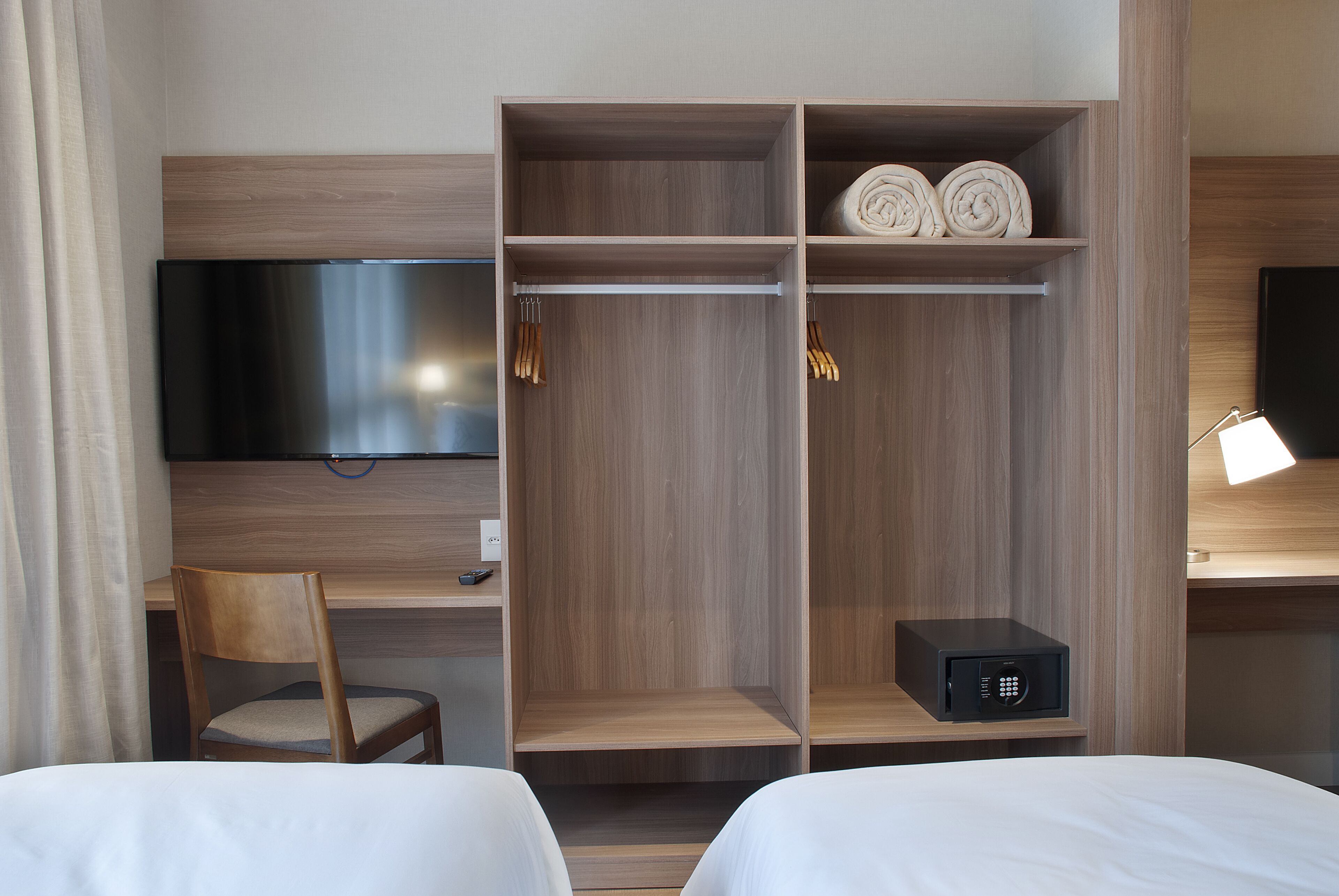A hotel room with a bed, a desk, a TV, a wardrobe, and a bedside table with a lamp.
