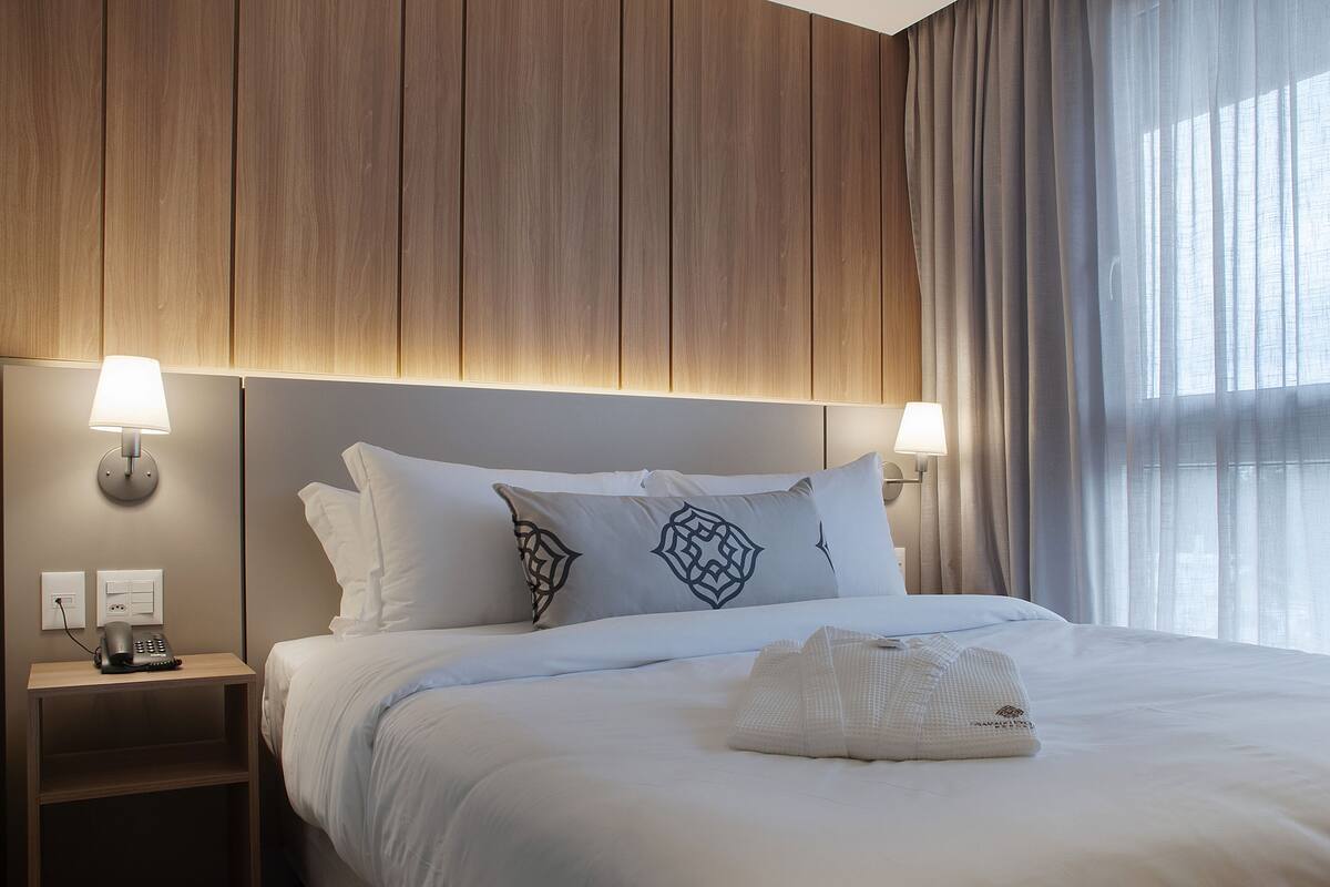 A neatly made bed with white linens and decorative pillows, a bedside table with a lamp, and a window with sheer curtains.