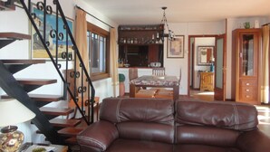 A living room with a brown leather sofa, a wooden dining table, and a staircase with black railings. - Santet CostaBravaSi - Pool, BBQ, garden, wifi (Calonge)