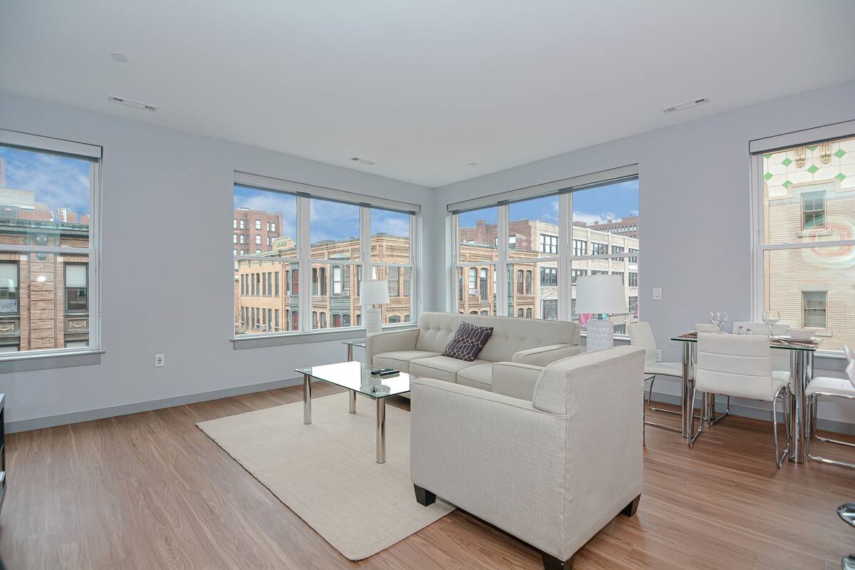 A modern living room with a sofa, coffee table, and dining area. Large windows offer views of buildings and a cityscape.