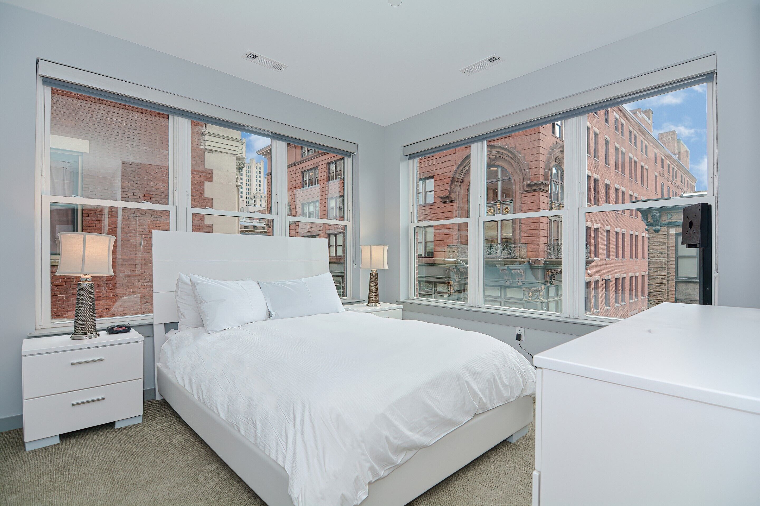 A modern bedroom with a large bed, two bedside tables with lamps, and a view of a brick building through large windows.