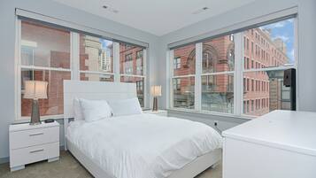 A modern bedroom with a large bed, two bedside tables with lamps, and a view of a brick building through large windows.