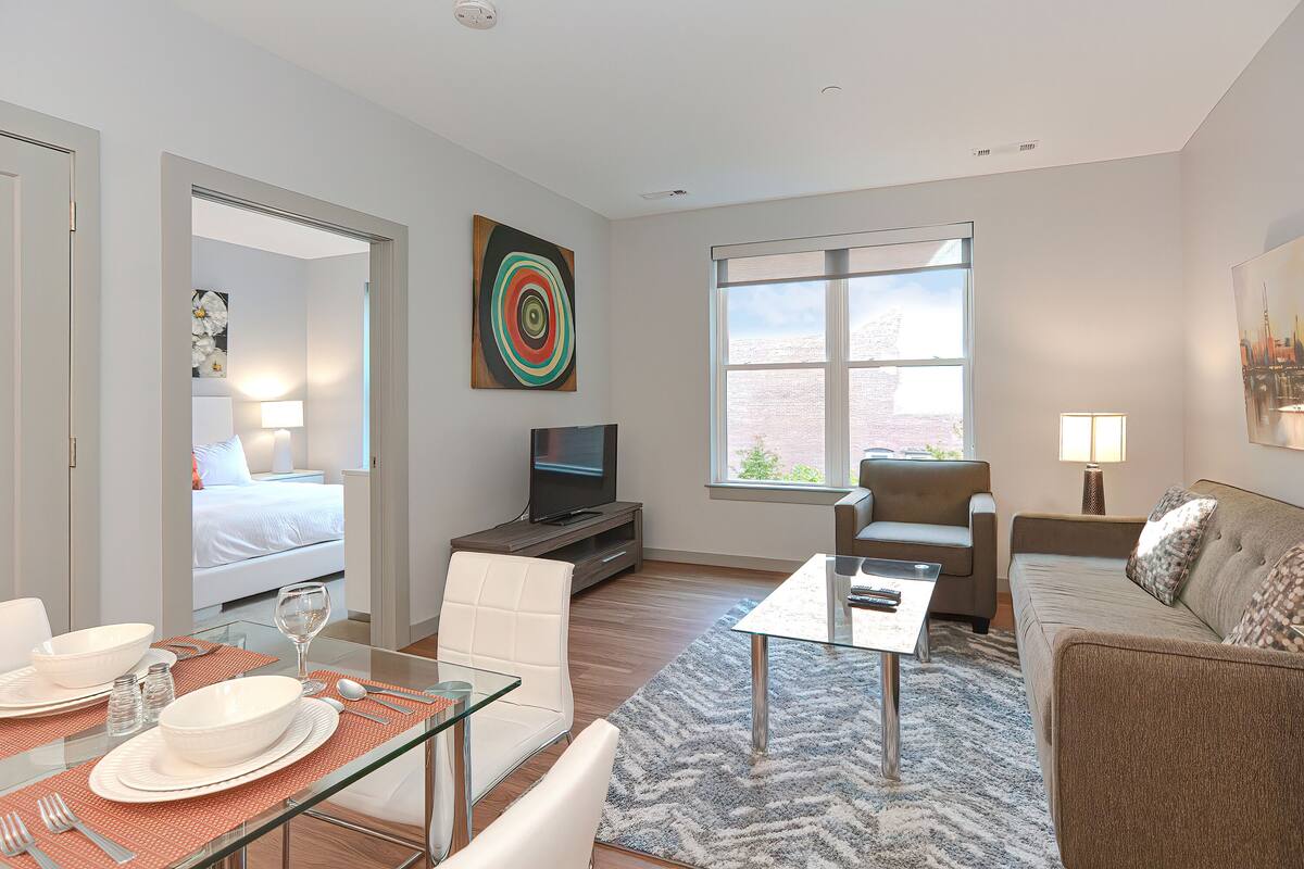 A modern hotel room with a dining area, a sofa, a TV, and a large window.
