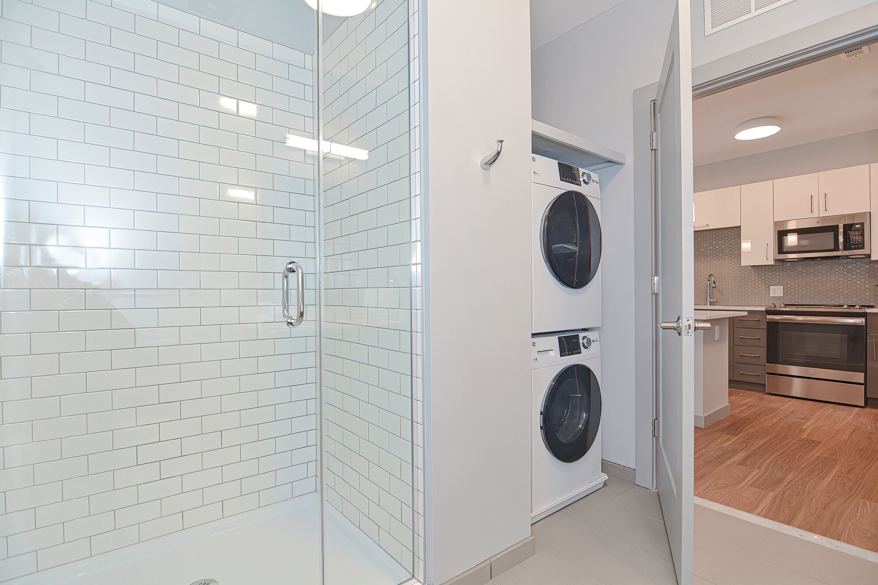 A modern laundry room with two washing machines, a glass shower enclosure, and a kitchen area with stainless steel appliances.