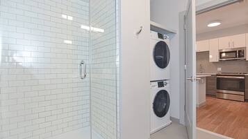 A modern laundry room with two washing machines, a glass shower enclosure, and a kitchen area with stainless steel appliances.