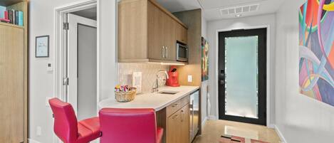 A modern kitchen with wooden cabinets, a microwave, and a red coffee maker. There are two red bar stools and a colorful abstract painting on the wall.