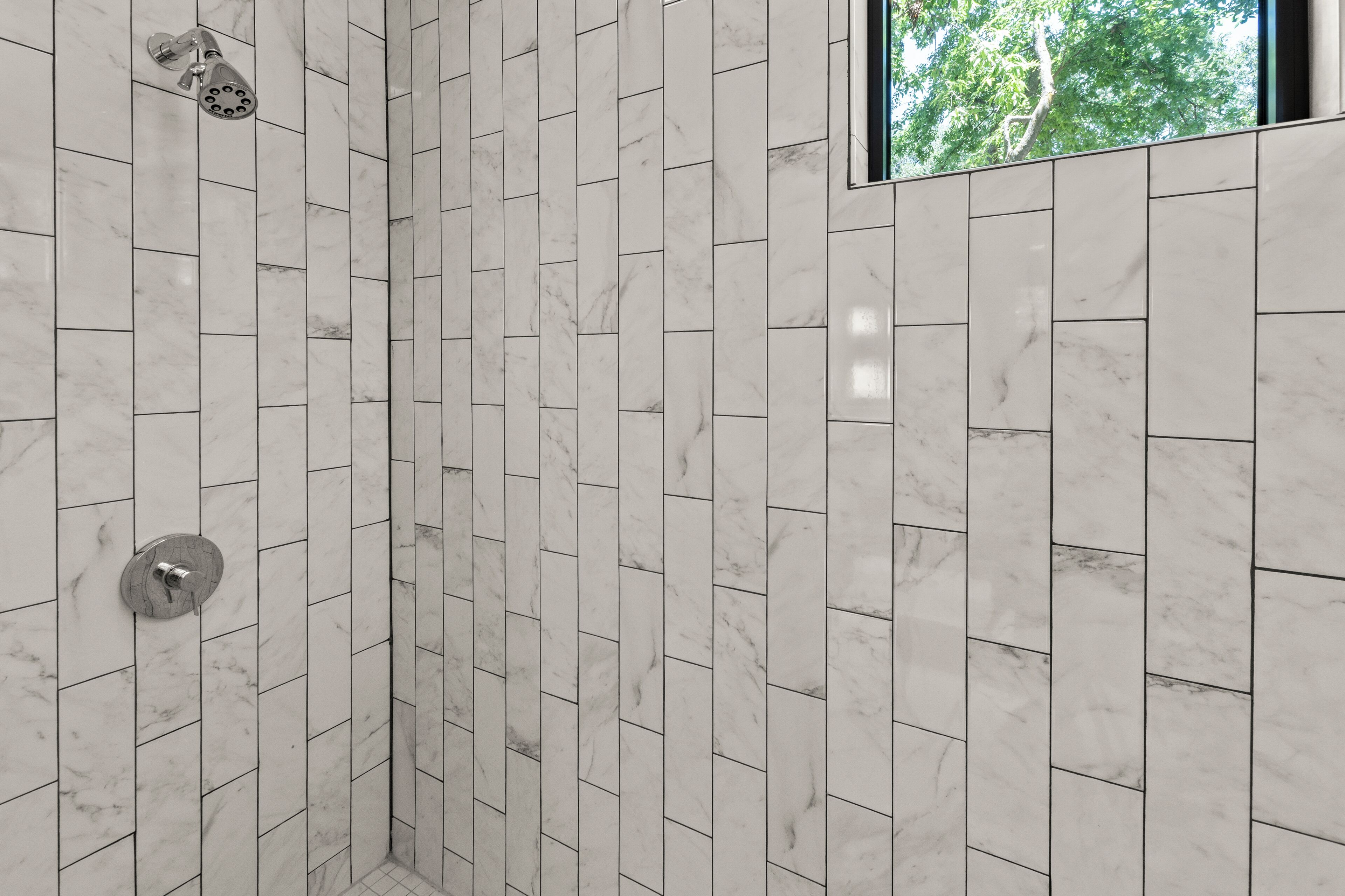 Une douche dont le carrelage est disposé en motif chevrons et une fenêtre donnant sur les arbres.
