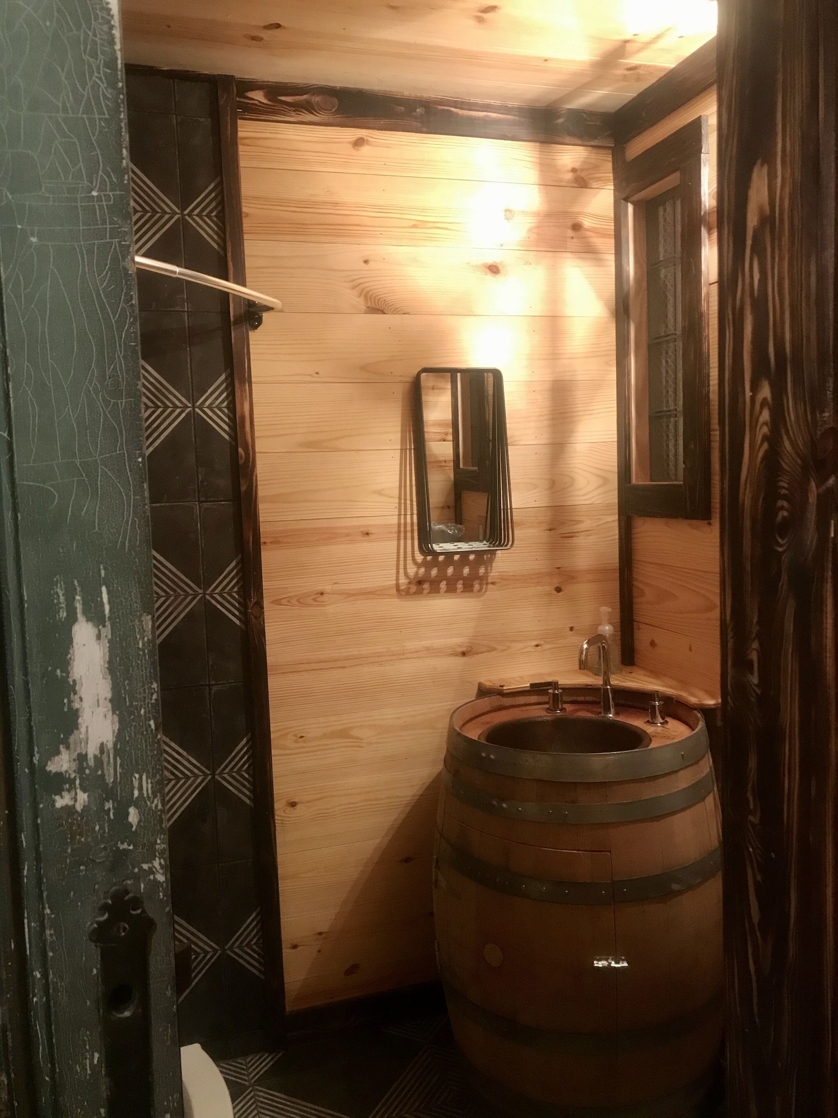A wooden interior with a wooden door, a mirror, a sink with a wooden barrel, and a patterned wall.
