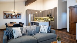 A modern living room with a gray sofa, a dining table with chairs, and a kitchen area with gray cabinets and stainless steel appliances.