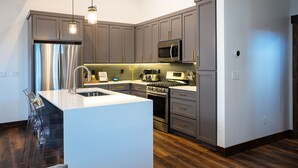 A modern kitchen with grey cabinets, stainless steel appliances, a central island with a sink, and a wooden floor.