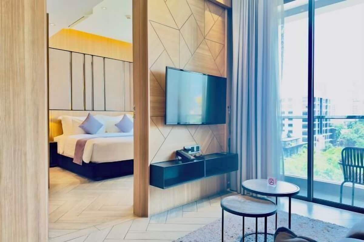 A modern hotel room with a bed, a flat-screen TV, a small table, and a large window offering a view of buildings and greenery.