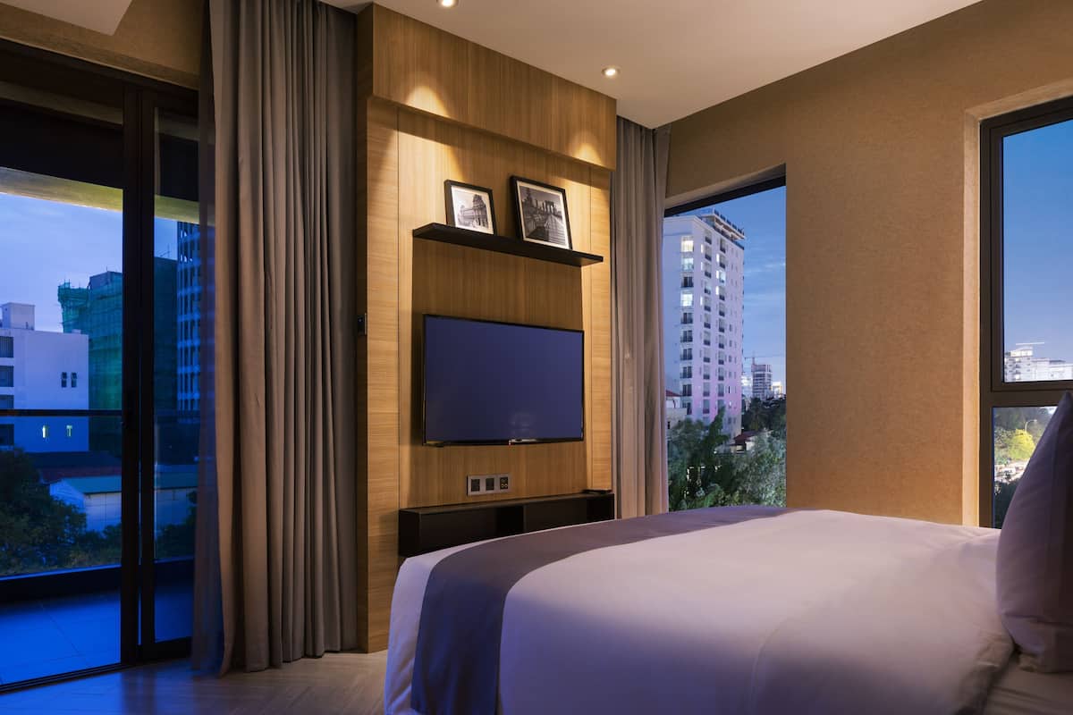 A modern hotel room with a large bed, a flat-screen TV mounted on a wooden panel, a sliding glass door to the outside, and a cityscape view.