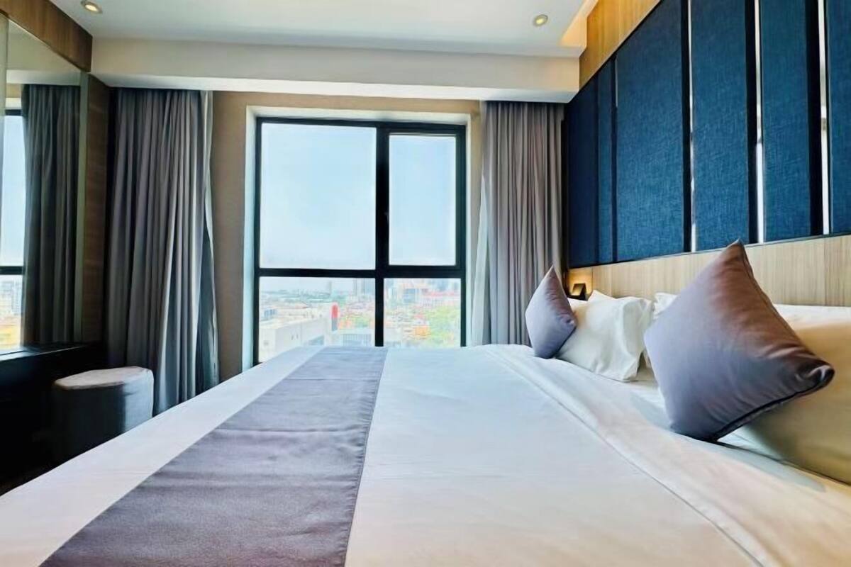 A hotel room with a large bed, a view through a window, and a cityscape.