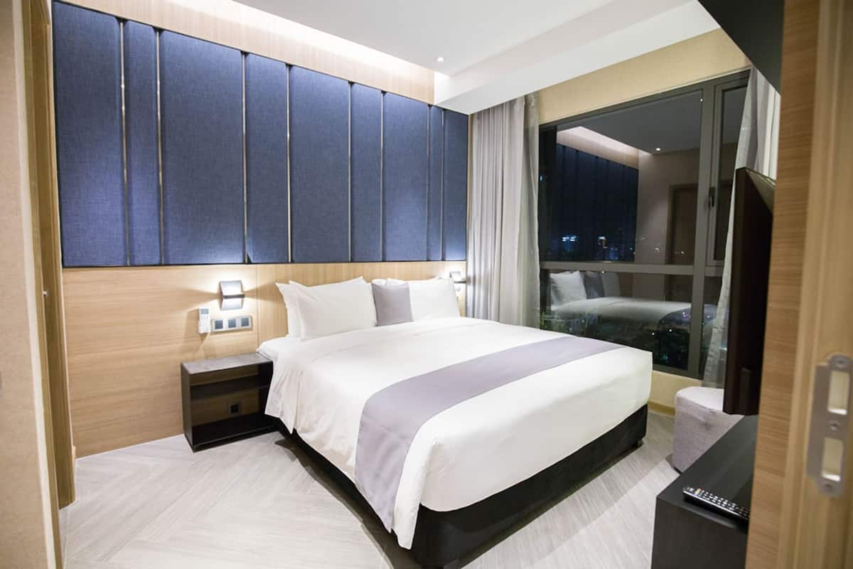 A modern hotel room with a large bed, a nightstand, a TV, and a view of the city at night.