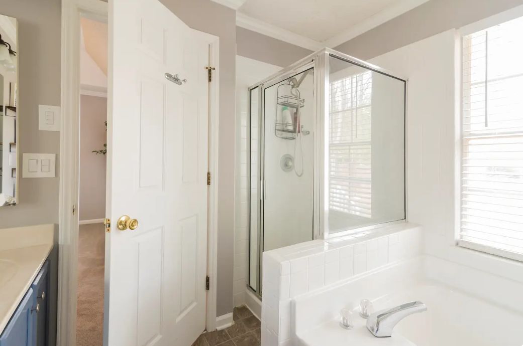 A bathroom with a glass-enclosed shower, a white sink, and a window with blinds.