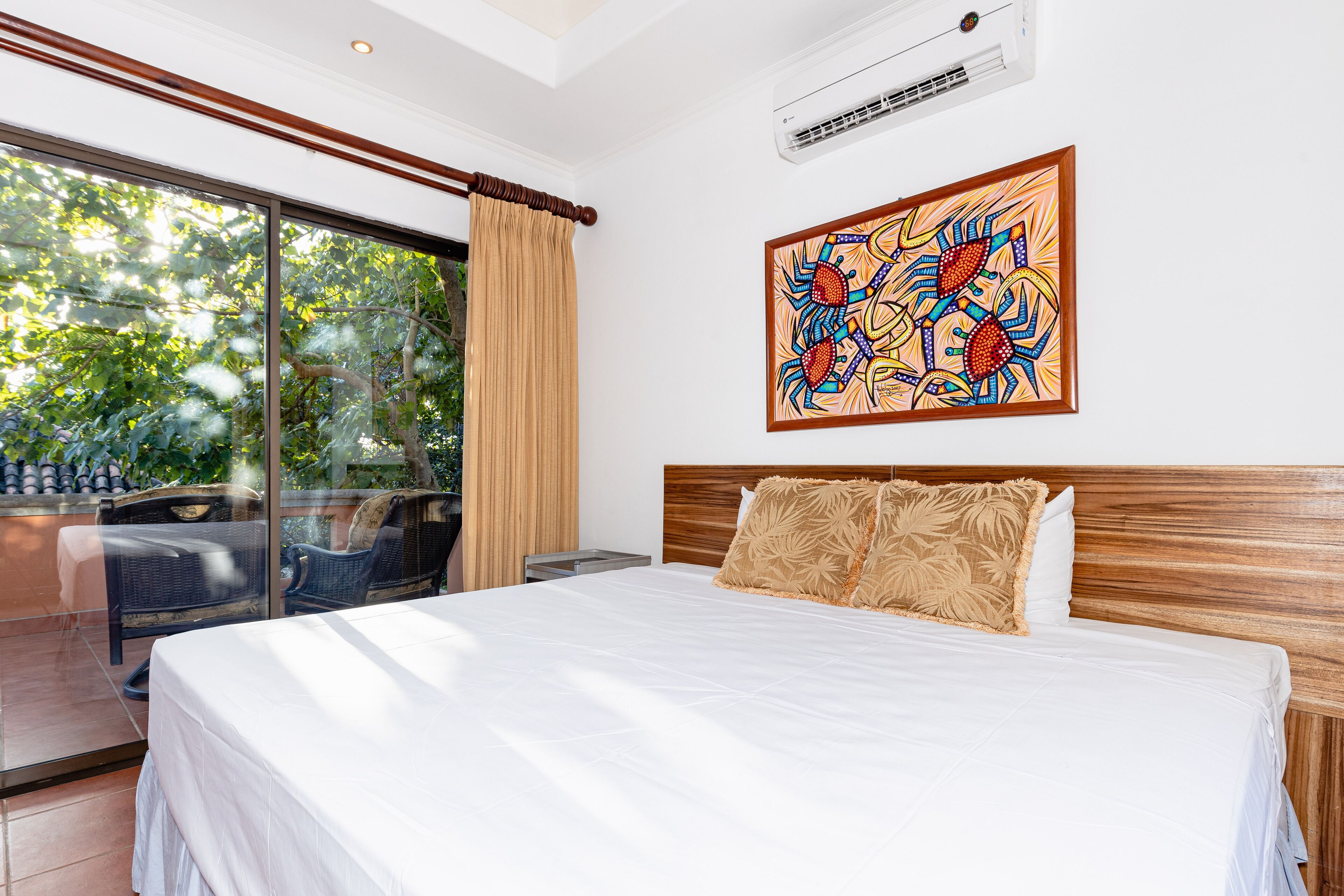 A hotel room with a large bed, a wooden headboard, a large window with a view of greenery, and a colorful artwork on the wall.