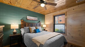 Quarto com teto de madeira, cama com colcha cinza, duas luminárias de cabeceira e janela com vista para um edifício.
