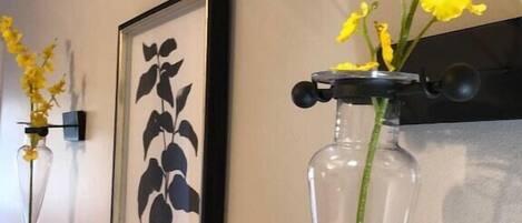 Un mur orné de deux reproductions botaniques encadrées et d’un vase transparent contenant des fleurs jaunes.