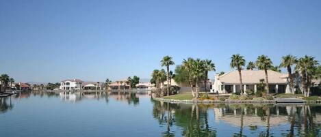 A serene lakeside view with multiple buildings, palm trees, and a clear blue sky.