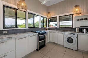 A modern kitchen with white cabinets, a built-in oven, a washing machine, and large windows offering views of the outdoors.