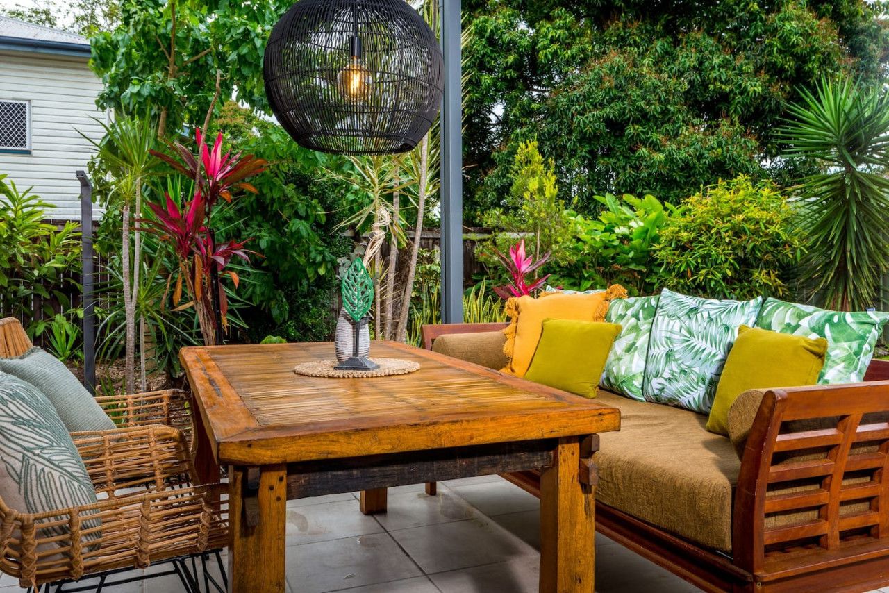 A wooden table with a woven chair, a sofa with cushions, and a hanging light fixture in a lush garden setting.