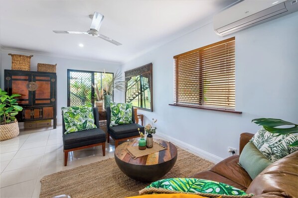 A living room with a ceiling fan, a wooden cabinet, two sofas with patterned cushions, a round coffee table, and a window with wooden blinds.
