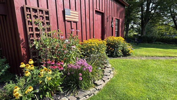 A red wooden building with a flag, surrounded by a well-maintained garden with various flowers and a stone border.